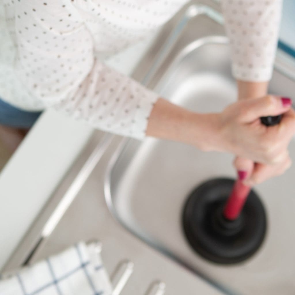 Someone using a plunger on a kitchen sink to help illustrate do I have healthy plumbing? and Is Draino Bad for Pipes?