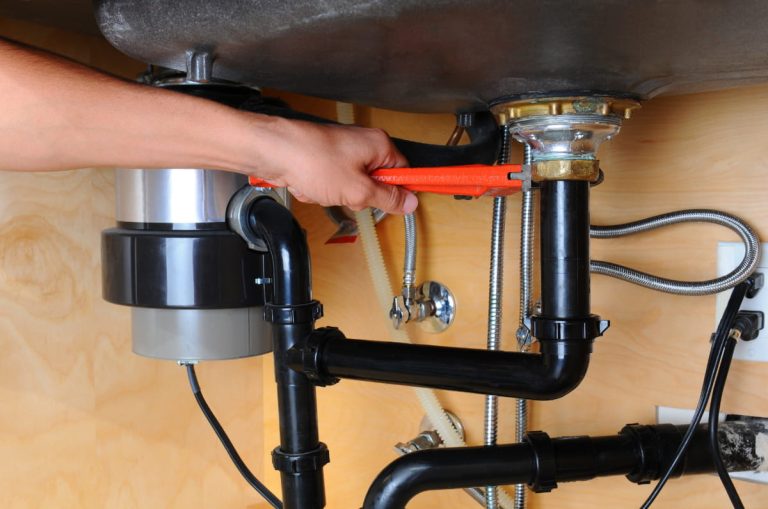 A plumber using a wrench to tighten a fitting beneath a kitchen sink to help illustrate A New Sink Means a New Cutout and Mounting Point during garbage disposal installation