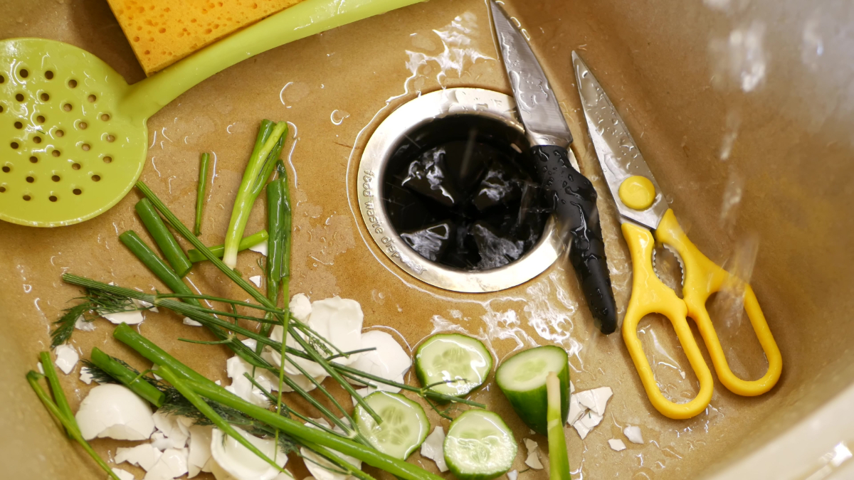 An image of kitchen sink with a garbage disposal and vegetable scraps, a spatula, knife, and scissors to help illustrate garbage disposal installation.